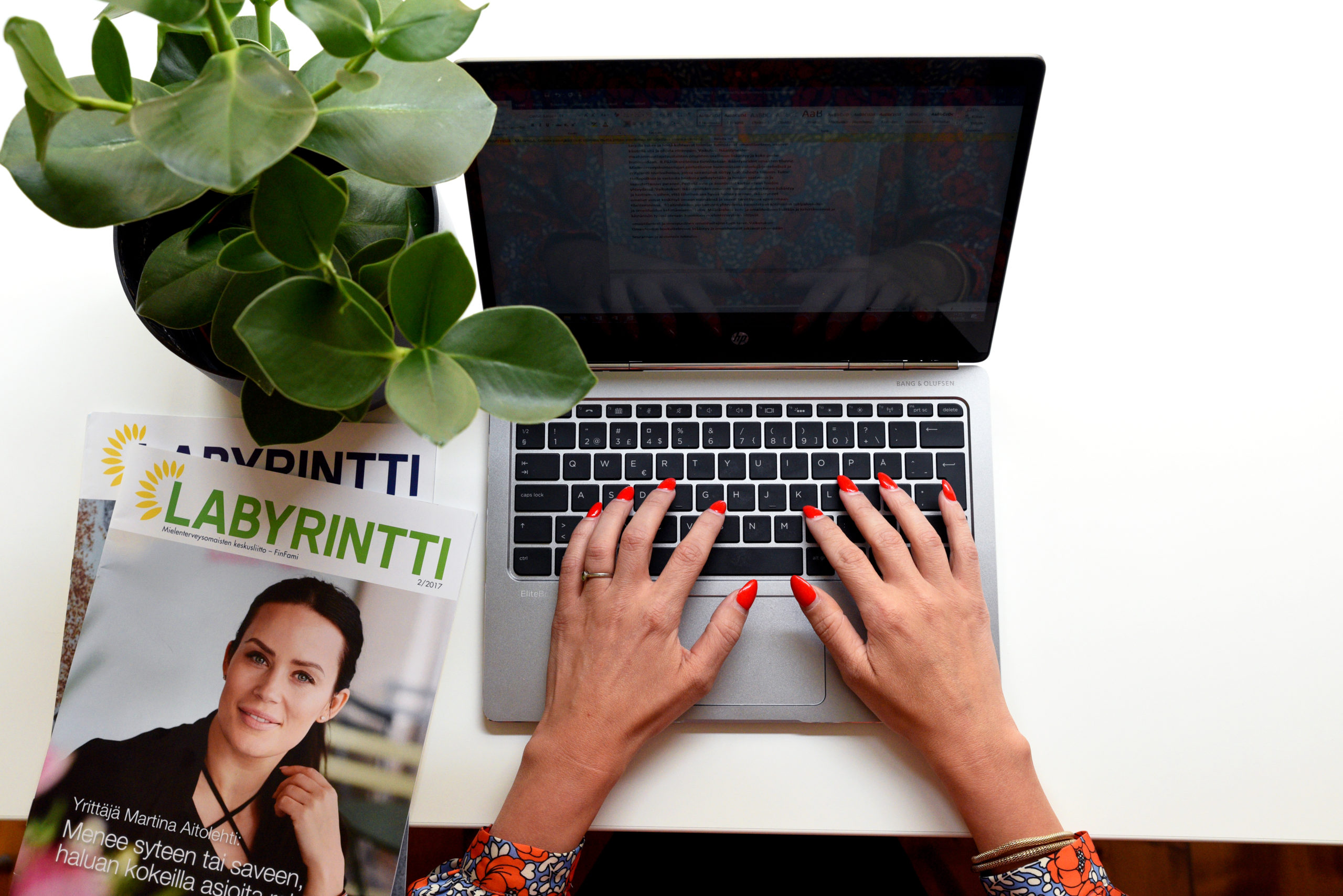 Käden kannettavan tietokoneen näppäimistöllä ja pöydällä huonekasvi sekä kaksi Labyrintti-lehteä.