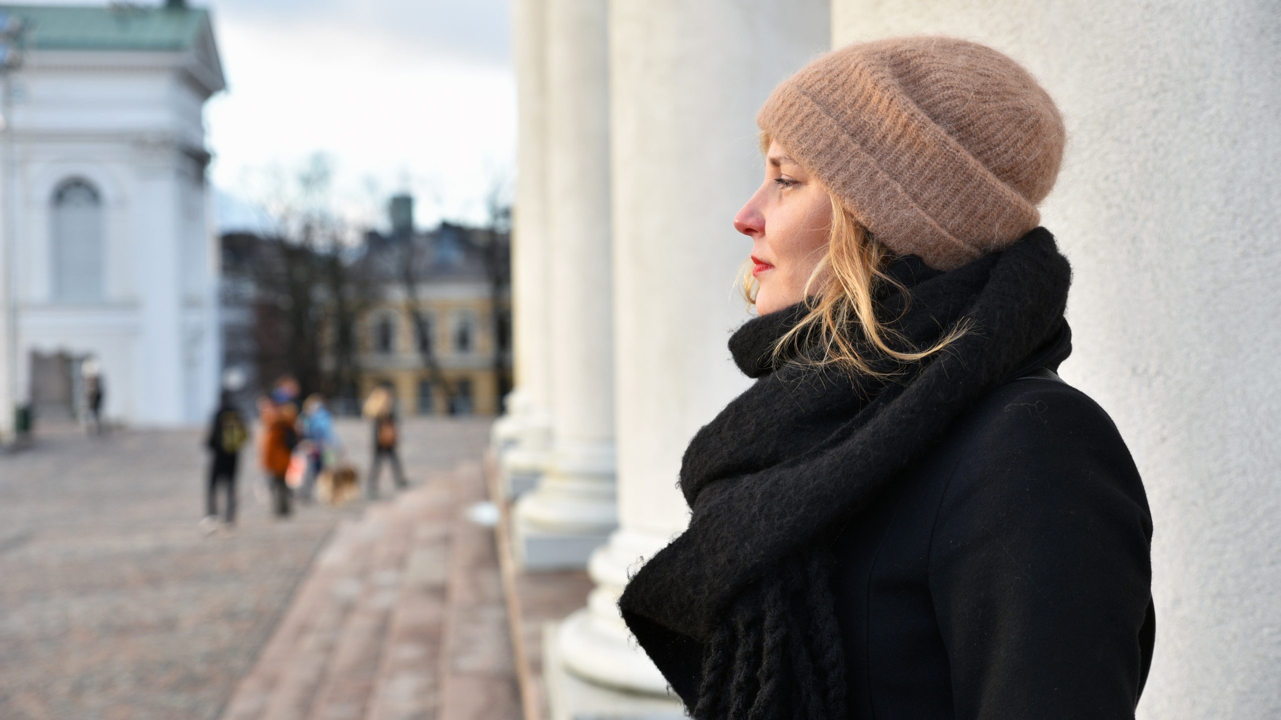 Naisen ulkovaatteissa, kasvot ja katse sivuun käännettyinä ja kaupunkimaisemaa sekä ihmisiä taustalla.