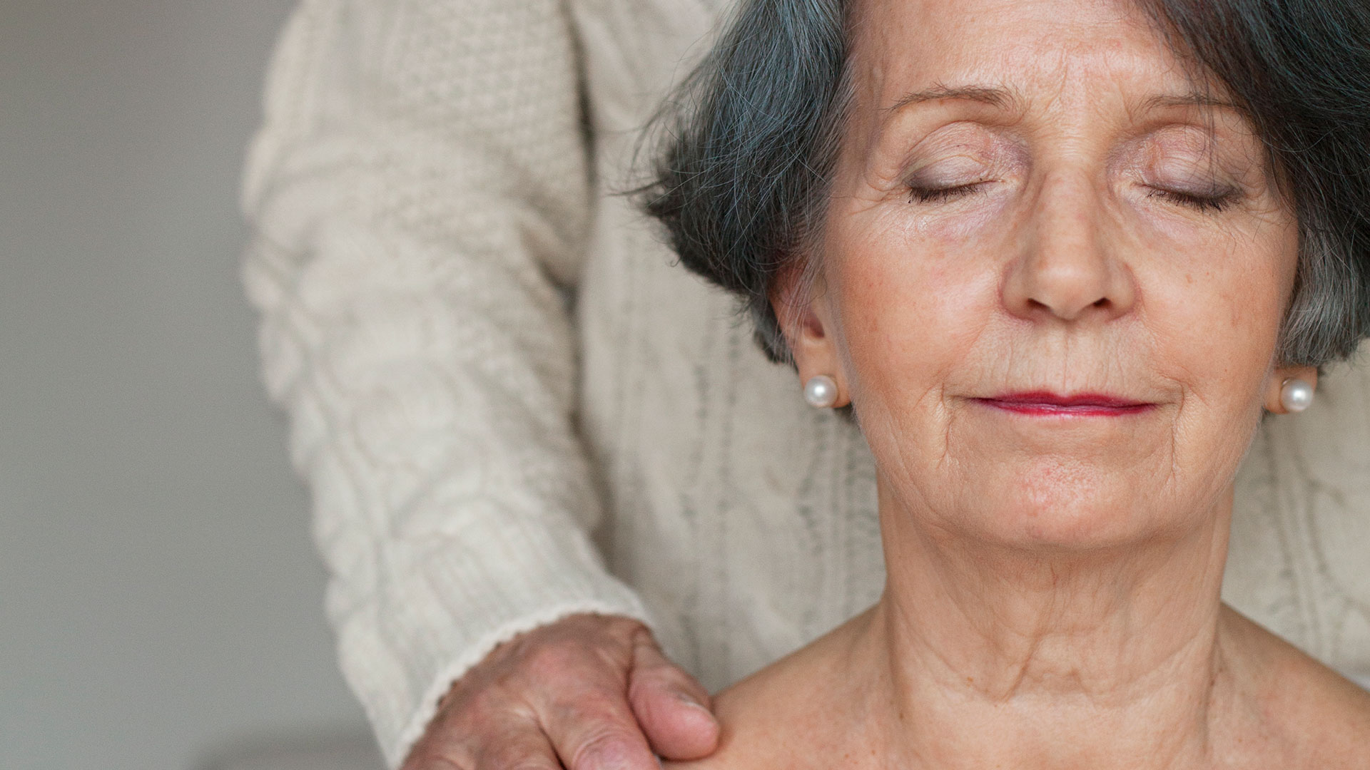 Nainen silmät kiinni kasvot kohti kameraa ja ihminen seisoo hänen takanaan, käsi hänen olkapäällään.